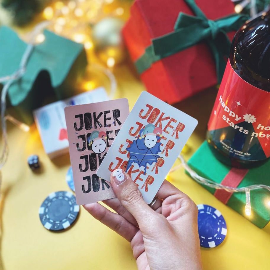 Hand holding Joker playing cards, Christmas presents, and bottle of wine on yellow background.
