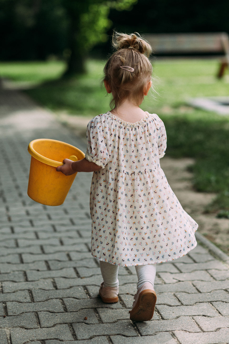 Mädchen mit hochgesteckten Haaren und hellem Kleidchen hat einen gelben Eimer in der Hand und läuft durch den Garten