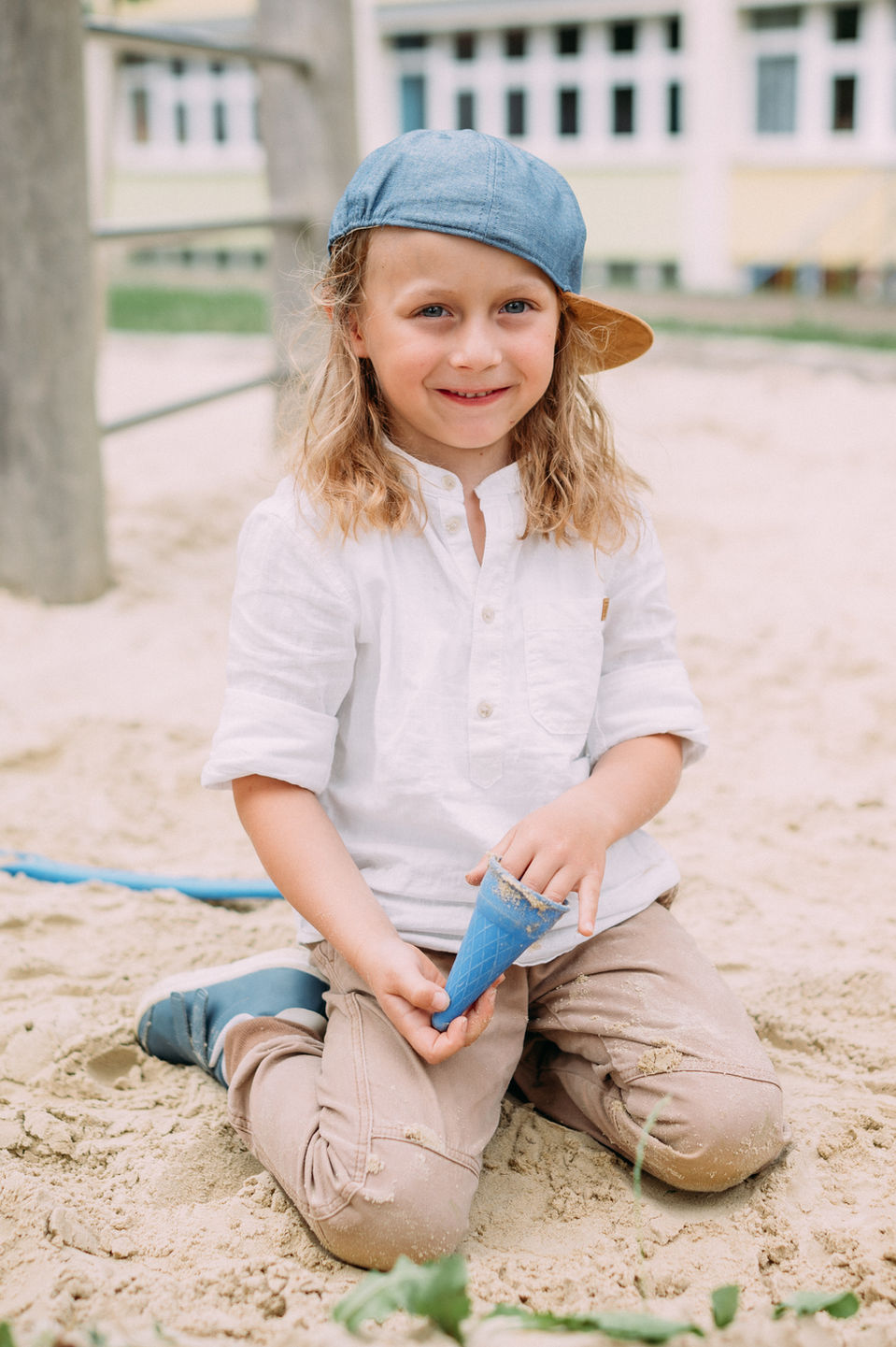 blonder Junge mit hellblauen Cap und beigefarbener Kleidung sitzt im Sand und lächelt freudig