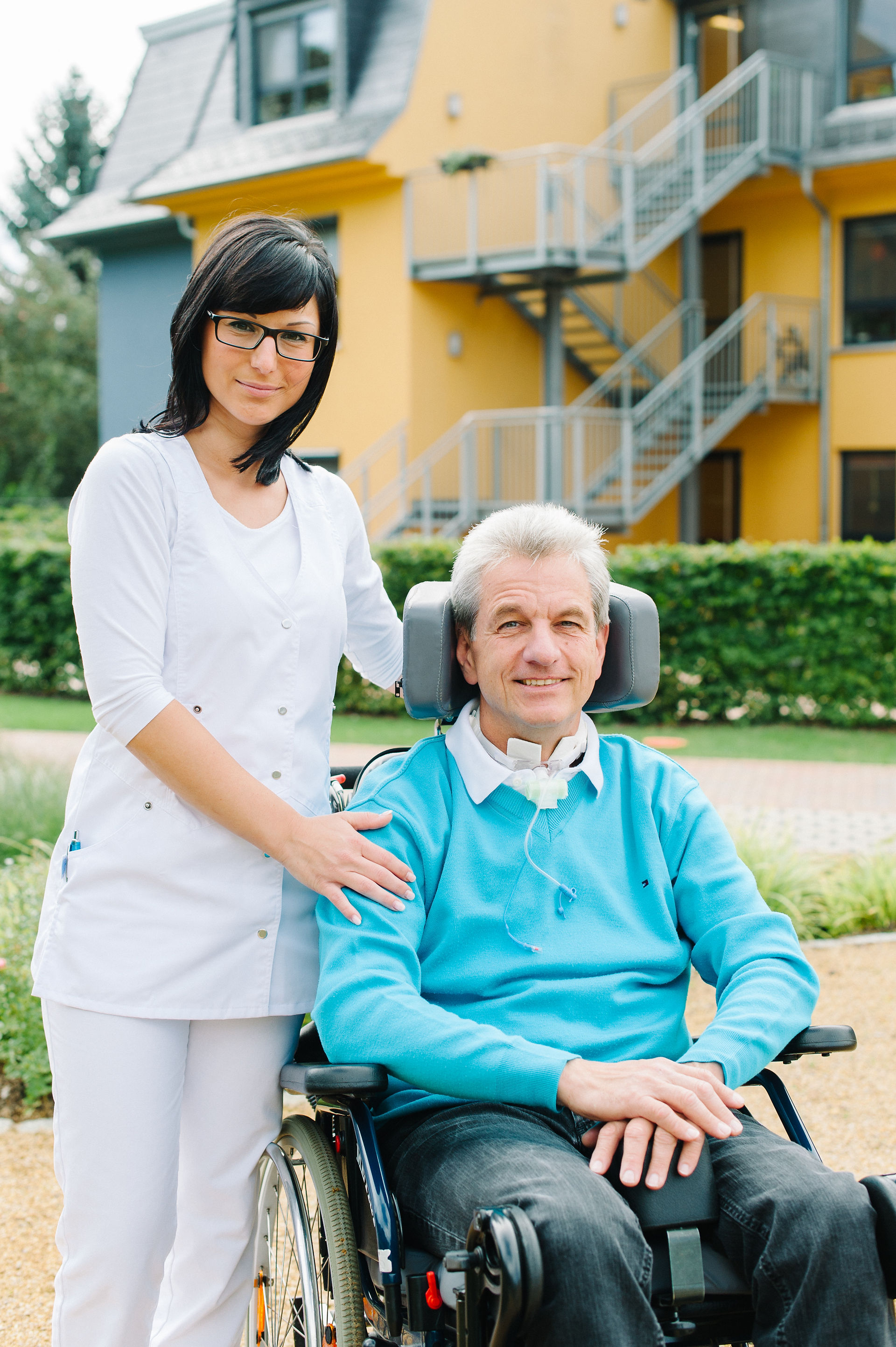 Krankenschwester steht draussen in der Sonne bei einem Mann mit grauen Haare im Rollstuhl