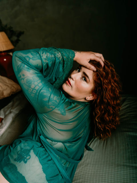 Artistic boudoir portrait of woman with curly auburn hair in translucent teal bodysuit, photographed on textured bedding with warm lamp lighting creating intimate atmosphere