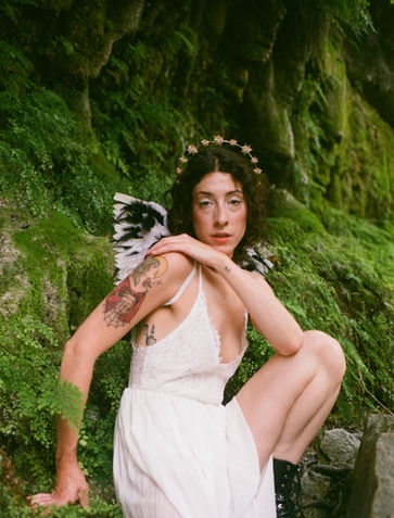 A bride with tattoos and a golden star crown sits among moss-covered rocks in a forest setting, wearing a white lace dress and black boots. The natural lighting and film grain create a moody, alternative bridal portrait.