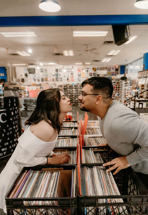 A couple leans over vinyl records in a vibrant store, playfully kissing. Bright lights and album covers fill the background, creating a joyful mood.