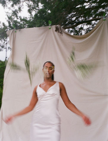 A bride in a white wedding dress stands with arms outstretched against a cream fabric backdrop, with intentional motion blur creating a dreamy, ethereal effect. Shot on film with natural lighting under tree canopy.