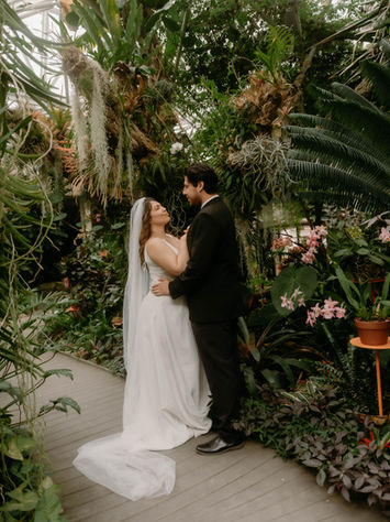 A bride and groom embrace in a greenhouse at the San Antonio Botanical Gardens. They are surrounded by plants.