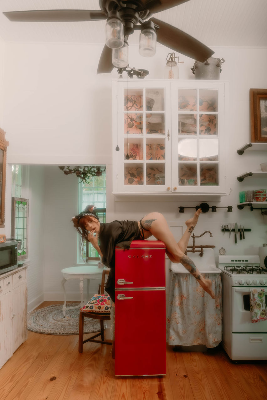 Editorial-style portrait in a vintage kitchen featuring a model posing on a red retro refrigerator, styled with tattoos, pin-up inspired fashion, and warm natural light. Creative lifestyle photography by an Austin, Texas photographer.