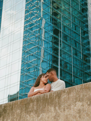 Romantic film photography of couple cuddling against concrete ledge with blue glass skyscraper reflection in Austin