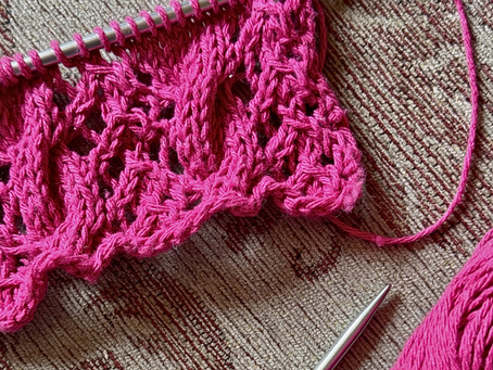 Pink lace knitting project with cables. Yarn ball nearby on a woven, earth-toned rug. Metal knitting needles 