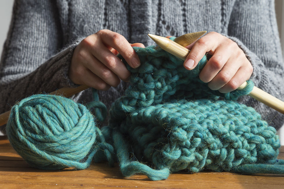 Hands knitting with large wooden needles, using thick teal yarn. A ball of yarn sits on a wooden table. Cozy and calm setting.