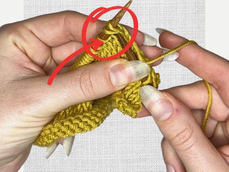 Hands knitting with yellow yarn and wooden needles, a red arrow pointing to a stitch. Neutral background texture.