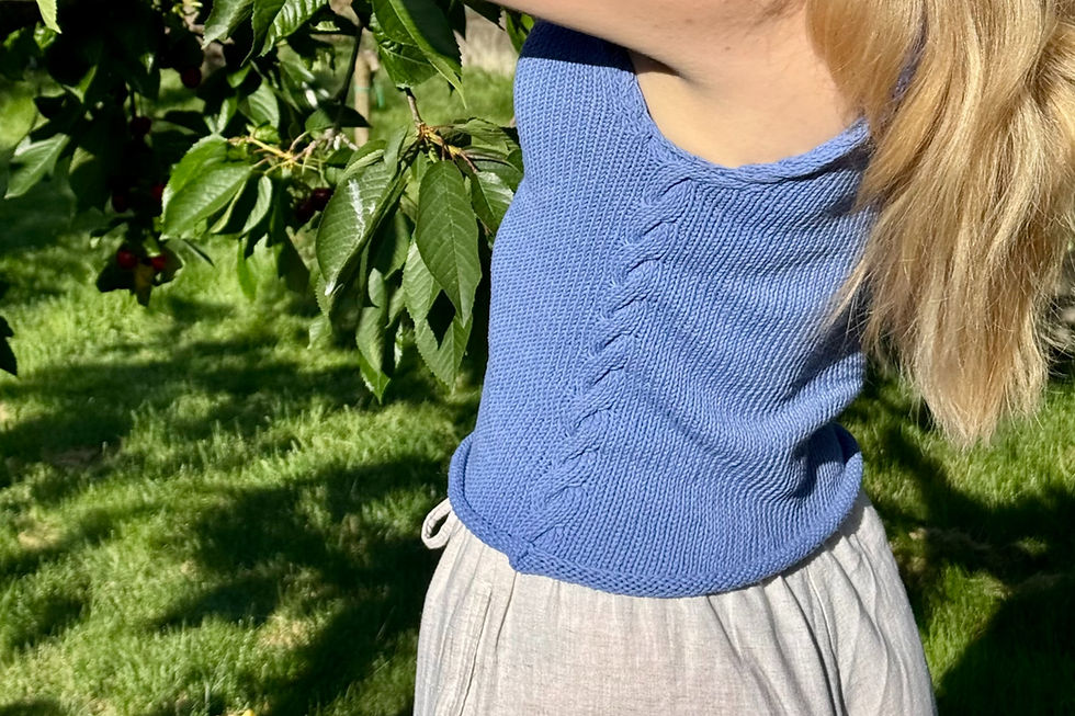 AnouskaKnits wearing a blue cable-knit top, The Olive Top, picking cherries in the sun, shadows casted behind her on the grass