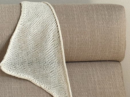 Cream, knitted Blueberry Bandana (pattern by AnouskaKnits) draped over a textured beige couch in a neutral-toned room. Soft lighting creates a calm, cozy atmosphere.