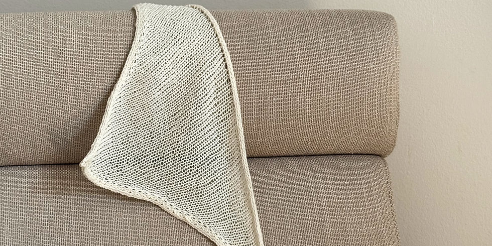 Cream, knitted Blueberry Bandana (pattern by AnouskaKnits) draped over a textured beige couch in a neutral-toned room. Soft lighting creates a calm, cozy atmosphere.