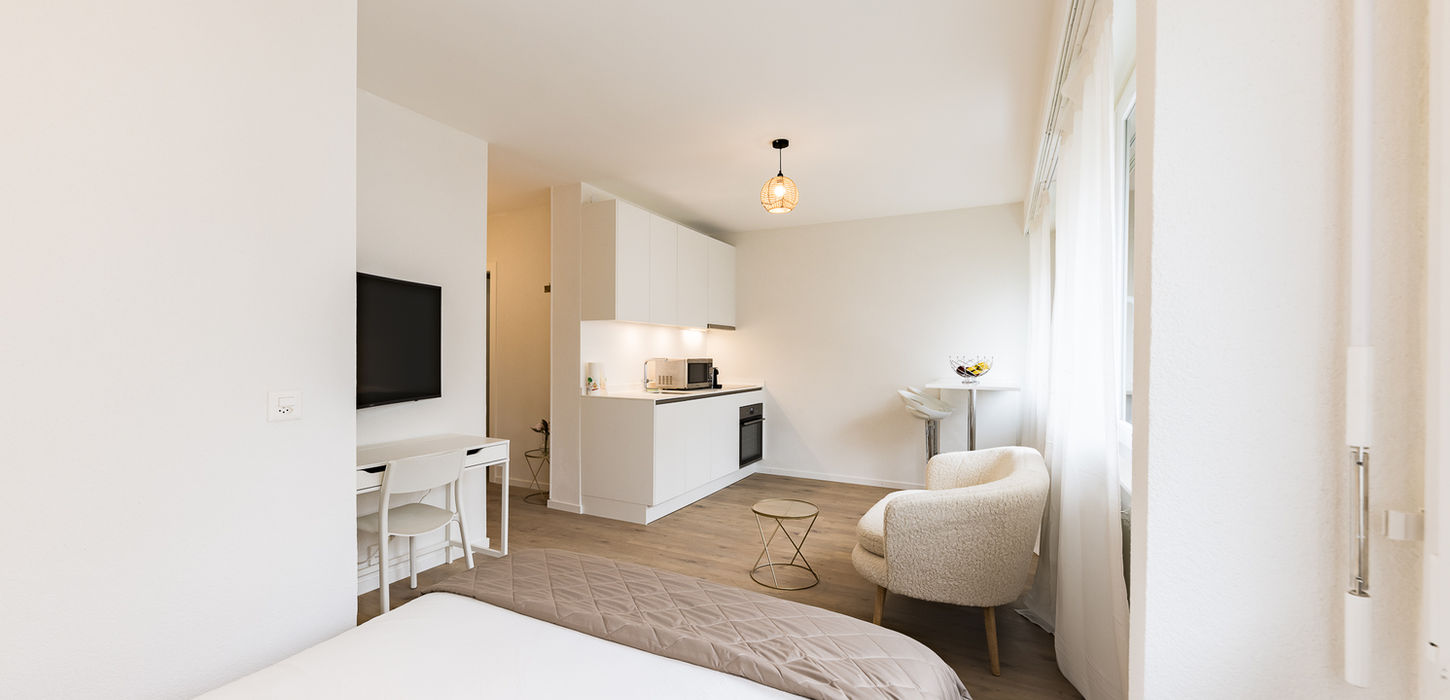 Modern studio apartment with white furniture, kitchen, and table; Swiss Flat Rental.