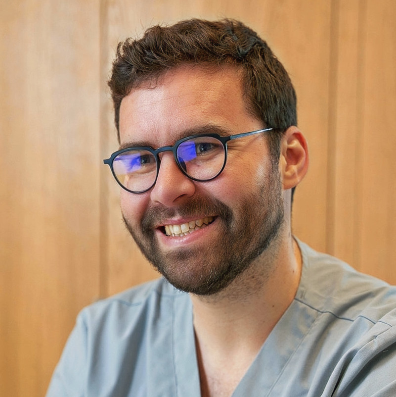 Smiling man in light blue scrubs and glasses looks professional.