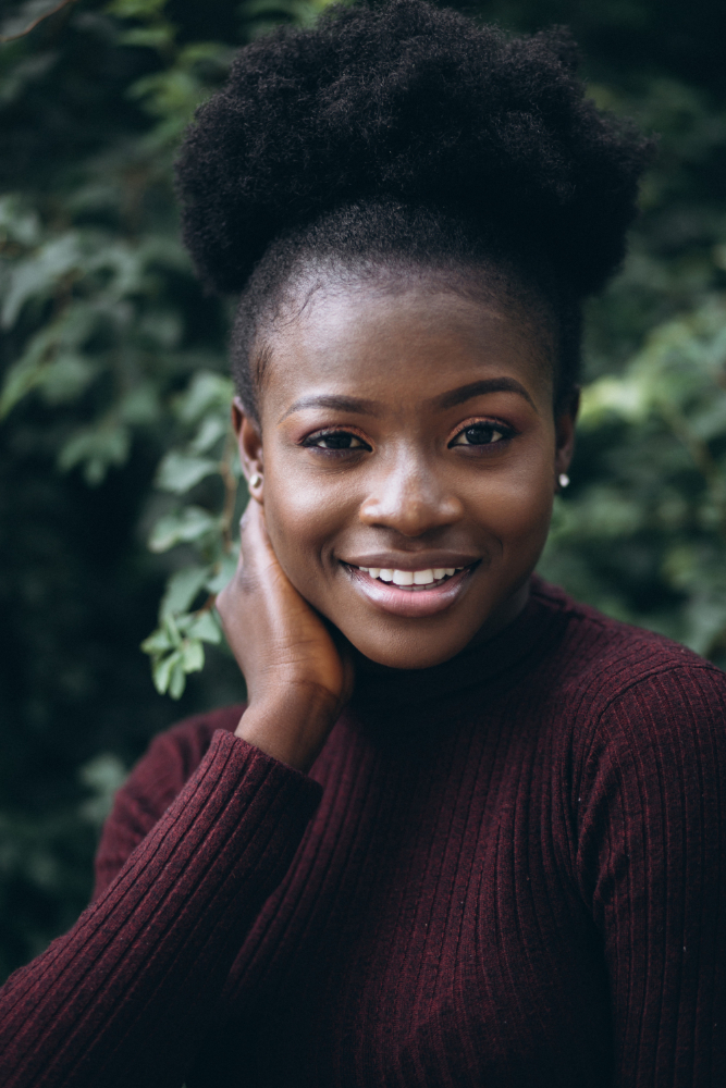 Smiling Black woman with afro, touching her neck against green foliage background.