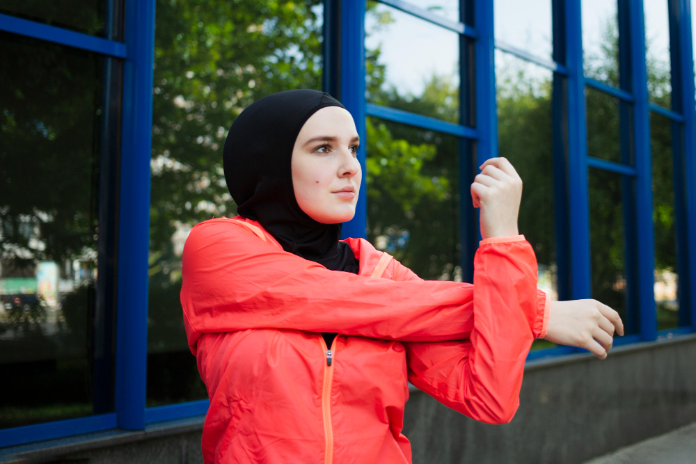 Young woman in hijab, stretching arms, wearing red jacket outdoors.