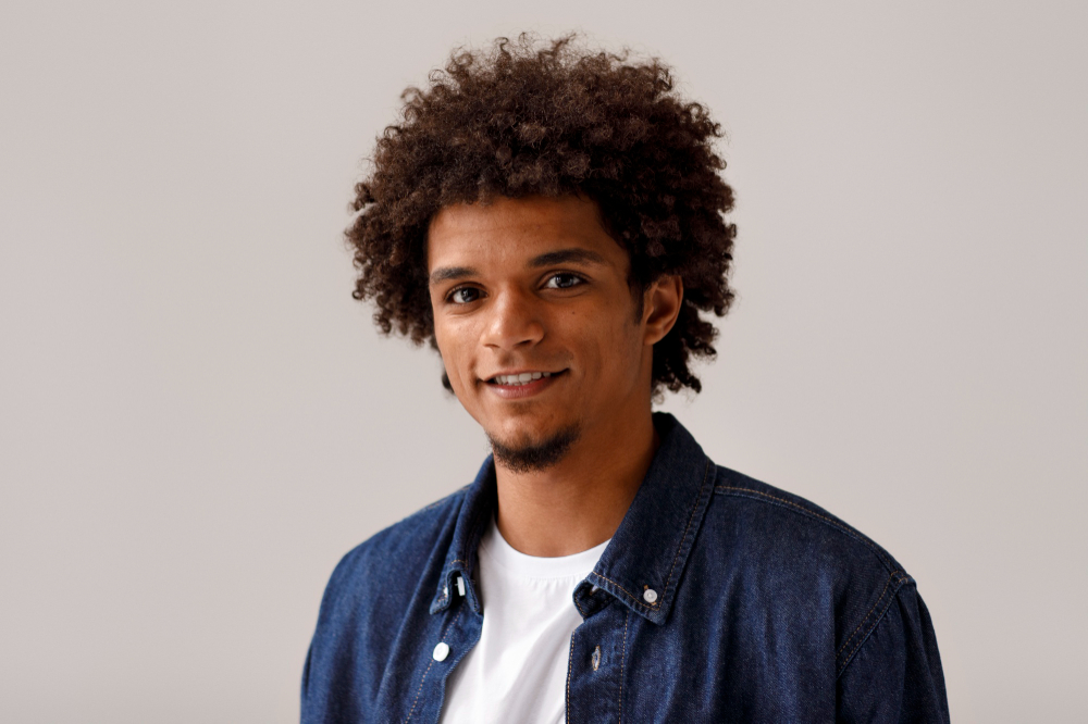 Smiling young man with curly hair and a denim jacket, looking at camera.