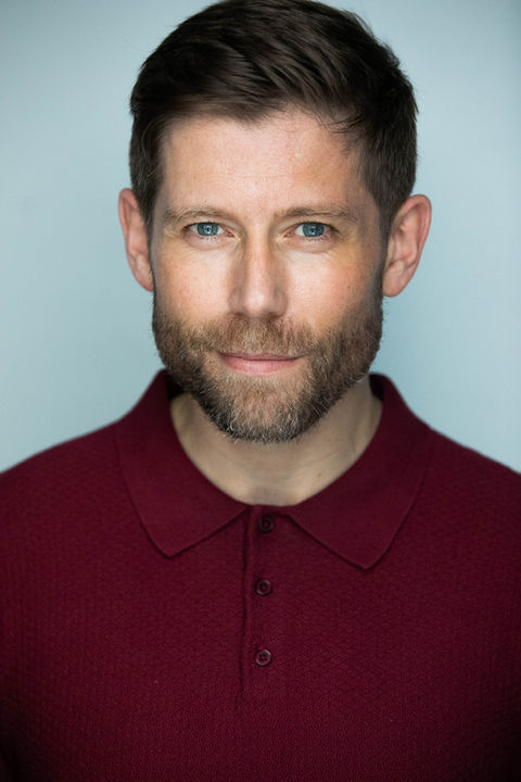 A Male actor in a burgundy collar shirt, he has a composed expression, there is a soft light grey background