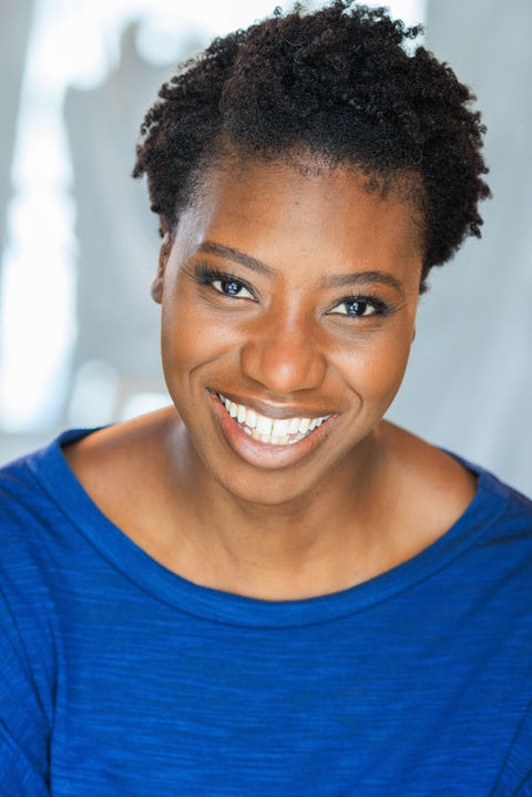 A Smiling actor with short curly hair in a bright blue top, she has a confident and engaging expression with a wide smile