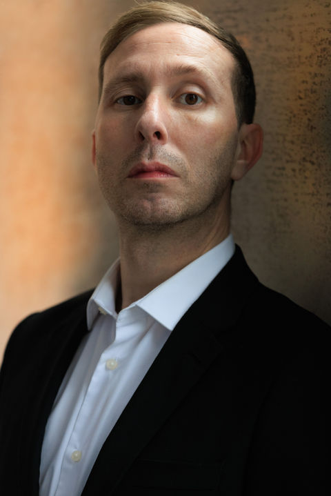 Male actor in a dark suit and white shirt, he has a serious and composed expression, there is a soft tan backdrop.