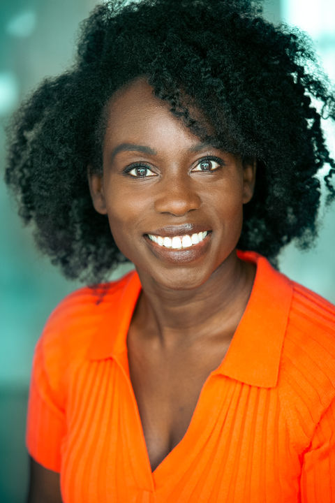 Actor with curly hair in orange shirt, she is smiling, and there is a soft blurred green background