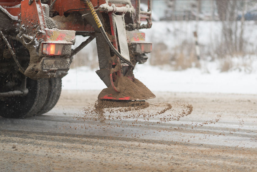 View behind a plow truck spreading de-icing salt and sand on the ice-covered asphalt road.