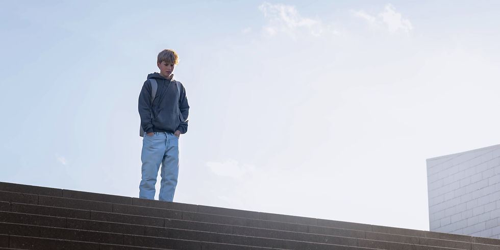 En dreng i mørk sweatshirt og lyse jeans står på en trappe og kigger ned. Klar himmel i baggrunden giver en rolig stemning.