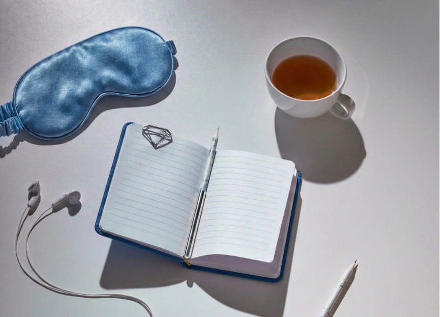 Open lined notebook with pen, tea cup, blue sleep mask, and earbuds on a white table in bright light.
