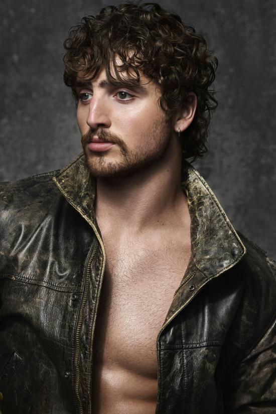 Curly-haired bearded man in a dark leather jacket, open chest, posing against a gray studio backdrop with a serious gaze.