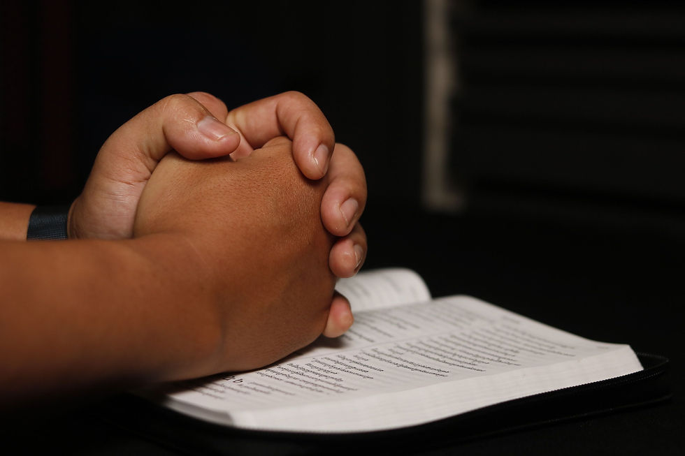 Clasped hands rest on an open book with blurred text in a dark setting, suggesting quiet prayer or reflection.