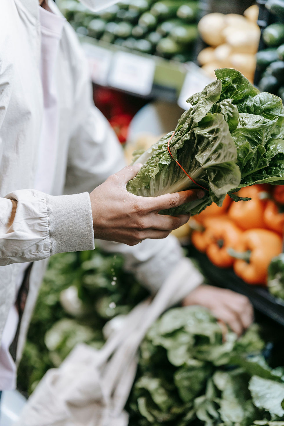 Hands holding a bundle of leafy greens in a grocery produce aisle, with blurred peppers and vegetables in the background