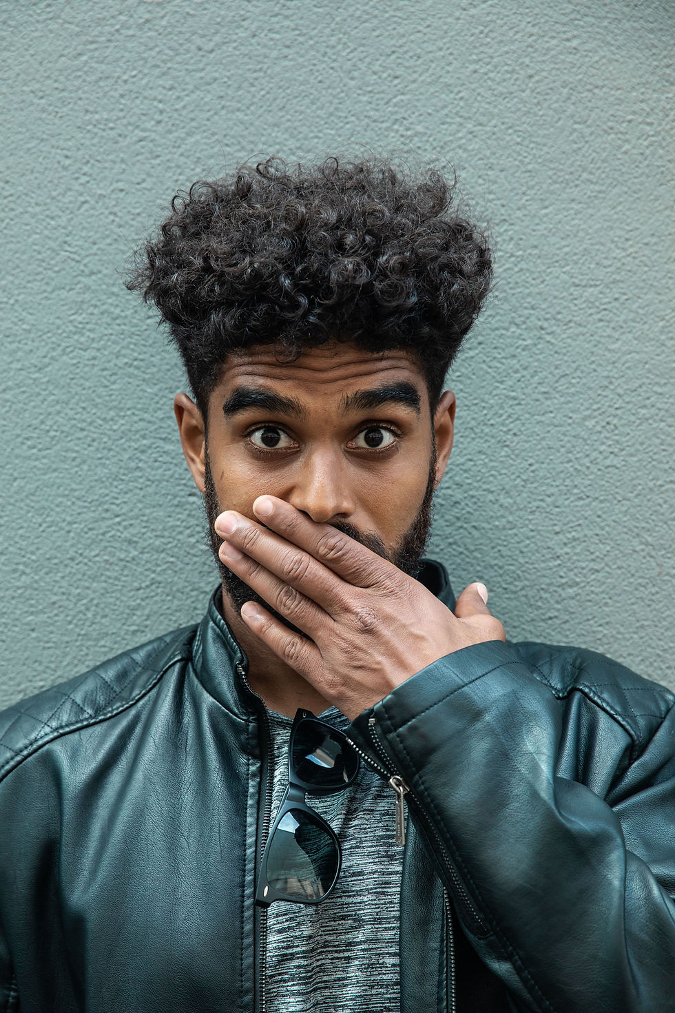 Young man in a black leather jacket covers his mouth in surprise before a textured gray wall, sunglasses hanging from his shirt.