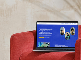 A laptop rests on a plush red chair, displaying a website about culturally responsive college readiness programs aimed at supporting first-generation students. The setting is cozy and inviting, featuring a neutral-toned wall and soft lighting.
