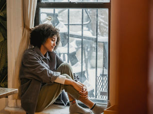 A woman sits by a window, eyes closed, looking slightly sad. Snow-covered scene outside. Warm, earthy indoor colors and leafy wallpaper.