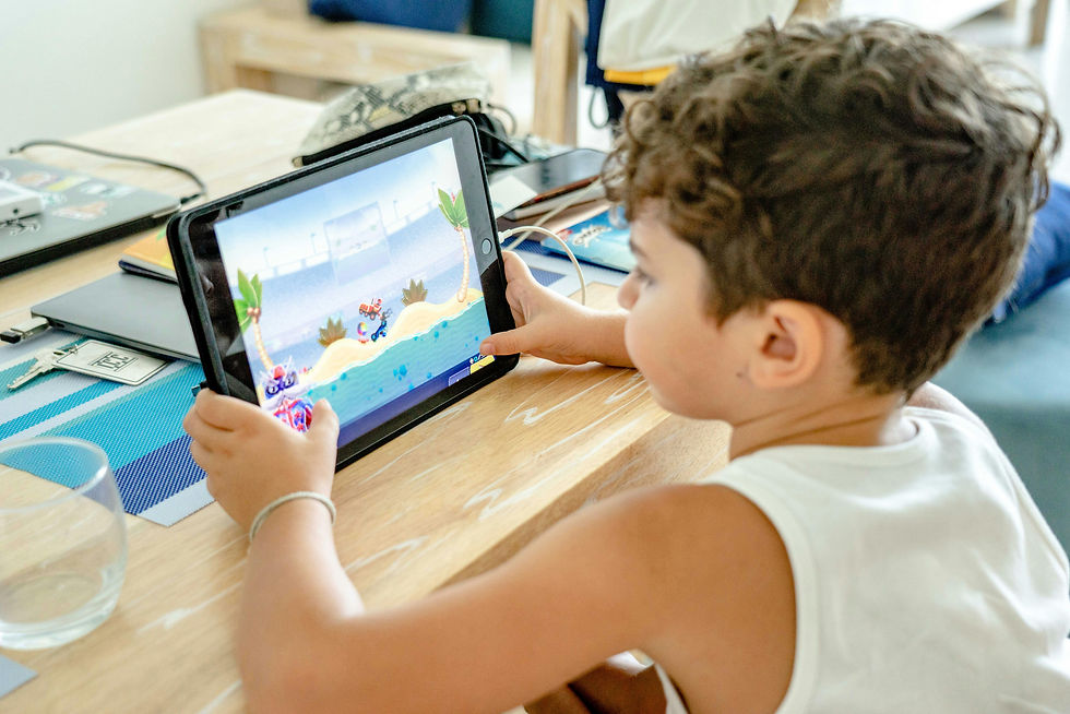 A young boy engrossed in a colorful game on a tablet, seated at a wooden table with notebooks and a glass nearby.