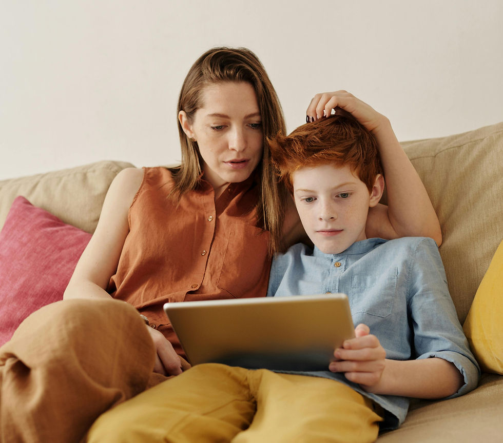 Woman and child sitting on a beige couch with colorful pillows, sharing a tablet. Woman in orange, child in blue; focused and relaxed mood.