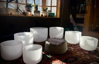 Eight white quartz crystal singing bowls on patterned rug, sunny window.