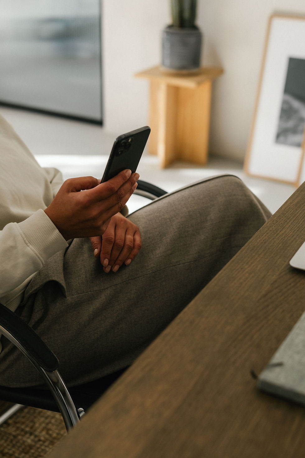 Person in beige shirt holding a smartphone, sitting at a wooden desk.
