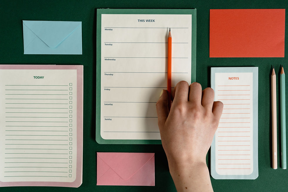Hand holding orange pencil over weekly planner on green desk, with TODAY and NOTES pads, envelopes, and two pencils nearby