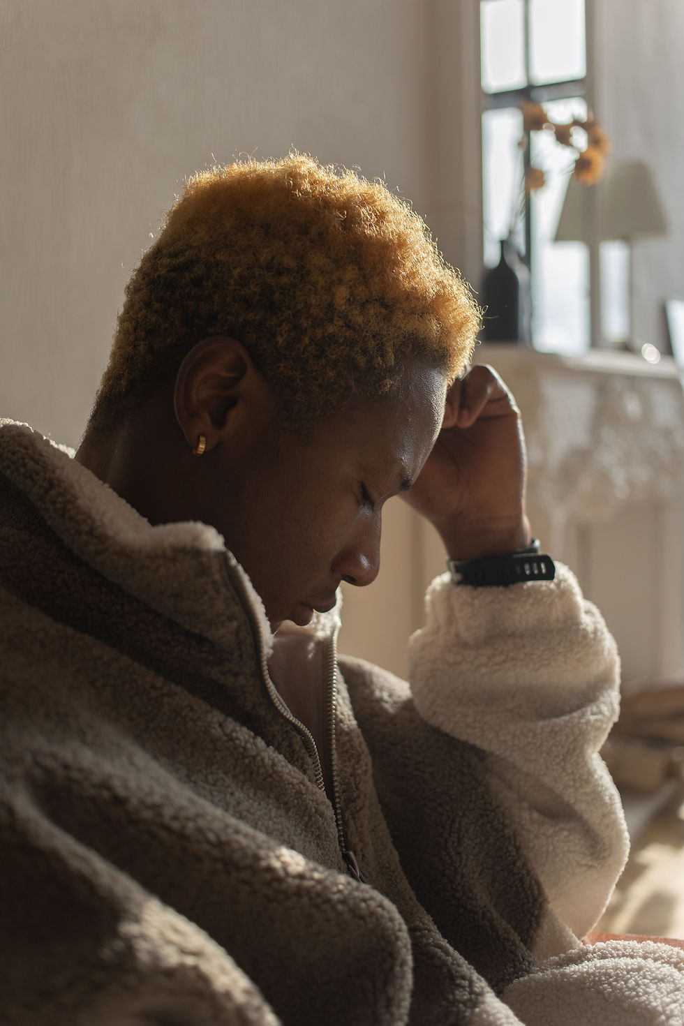 Profile of a pensive person in a tan fleece jacket, hand to forehead, seated in warm sunlight by a window with a lamp behind.