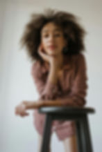 Young woman with curly hair leaning on a stool, chin on hand.