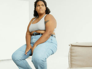 Person sitting on a stool in a white room, wearing a gray tank top and blue jeans. They appear thoughtful, with a calm expression.