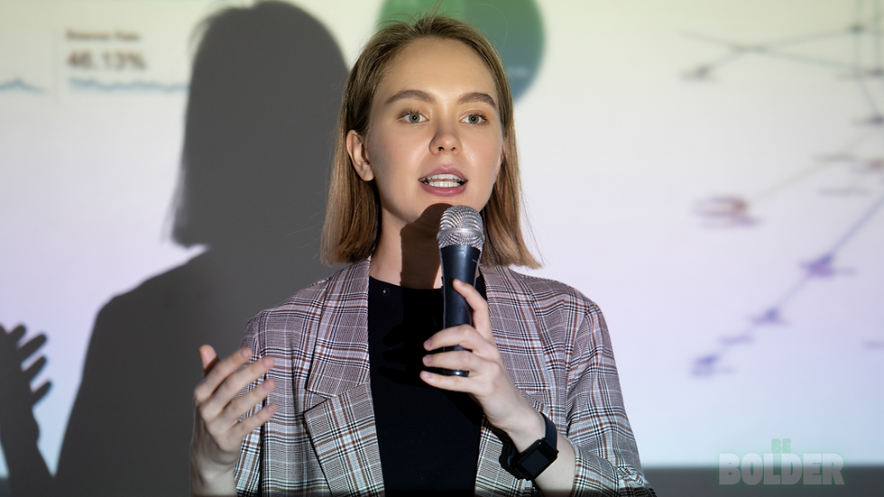 Woman presenting with a microphone in front of a projected graph. She wears a plaid blazer and a black shirt. Text on screen reads "BE BOLDER."