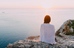 A person in a white shirt sits on a rocky cliff overlooking a calm ocean at sunset, creating a serene and contemplative mood.