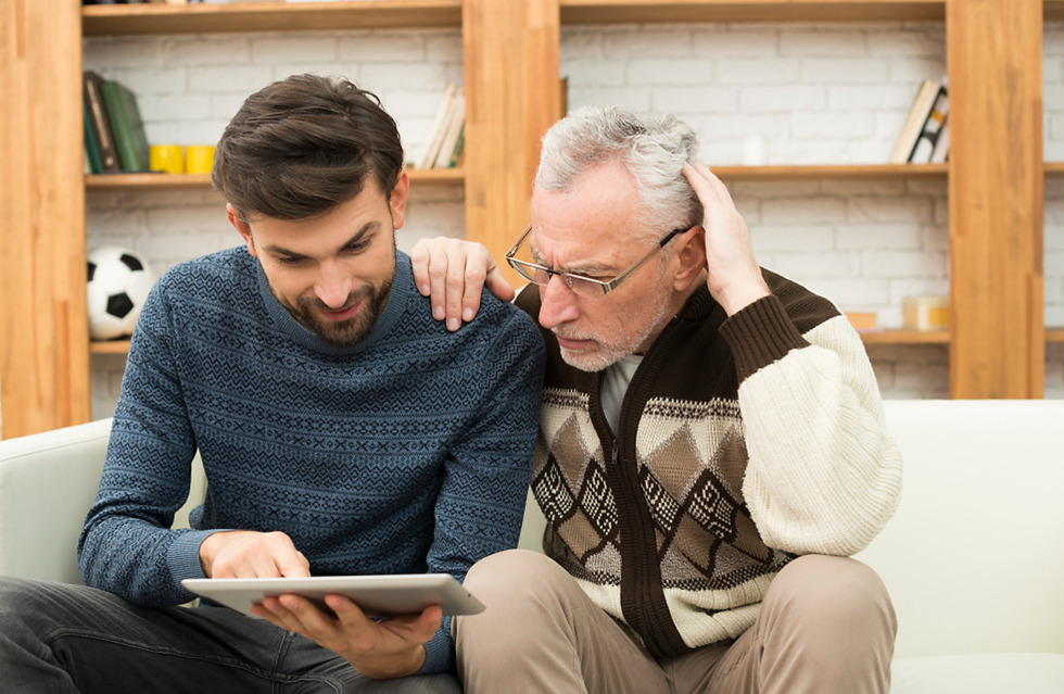 Elderly man and younger man sharing a tablet on a couch, surrounded by bookshelves. They're focused and engaged. Cozy setting.