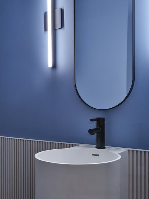 Blue washroom with ribbed wainscoting, rounded mirror, white pedestal sink, and vertical LED sconce.