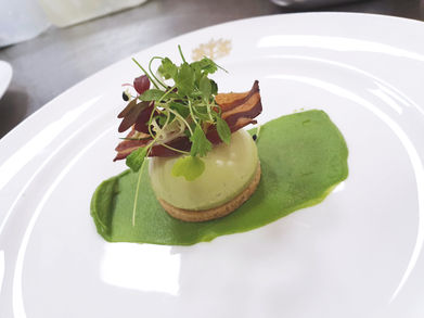 A savory broccoli and Montgomery cheddar cheesecake on a parmesan shortbread base, plated with broccoli purée, crispy bacon, and fresh microgreens on a white plate. A creative and elegant fine dining dessert.