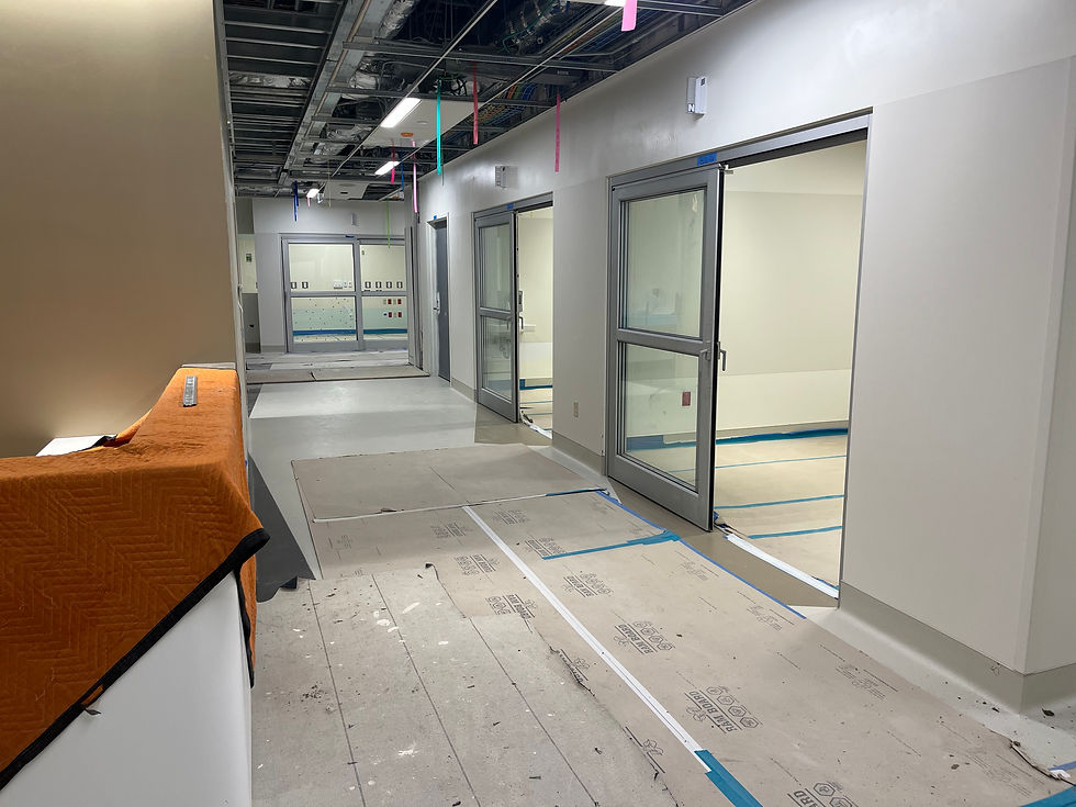 Hallway with finished beige walls, floors are covered with construction cardboard and blue painters tape, furniture is covered with orange protective construction blankets, ceiling has some panels but mostly exposed to the mechanical, electrical, and plumbing, multiple rooms with sliding glass and medal doors. room to the very back shows the headwall with electrical and medical gas boxes and finish plates