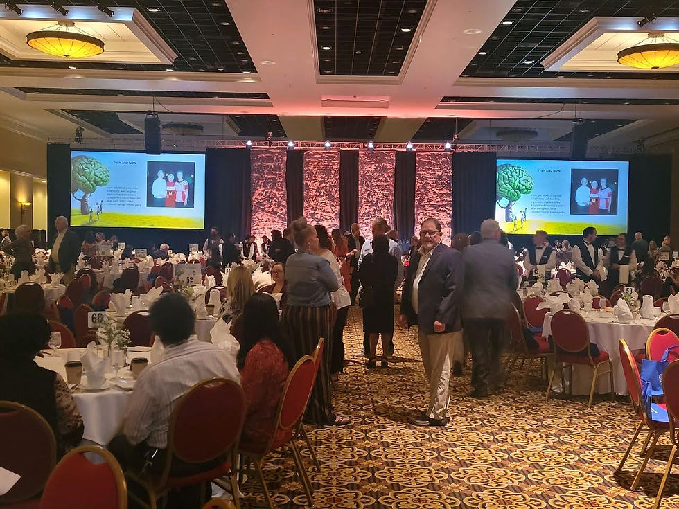 Attendees of the event gathered around networking or sitting at round tables with white table cloths and red and gold chairs, back ground is the stage with two screens on each side displaying text and a photo, middle of stage is a reddish brown background lite up with dark brown drapes, flooring is multi colored carpeting, ceiling is white and black with yellow paneled large round lighting fixtures.